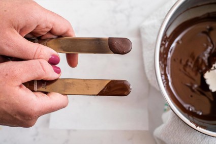 A baker holding two offset spatulas that are being used to test chocolate in temper