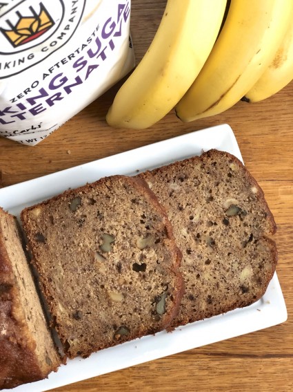 Banana bread made with Baking Sugar Alternative, sliced on a white plate with bananas in the background.