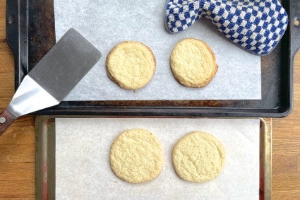 Four cookies on two baking sheets, one light and one dark, to show the difference in browning between cookies baked on a dark pan and those baked on a light pan.