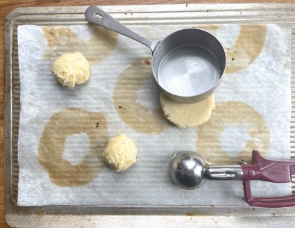 Three balls of Sweet Corn Cookies dough on a baking sheet with a scoop, one ball beinggently flattened with a dry measuring cup.