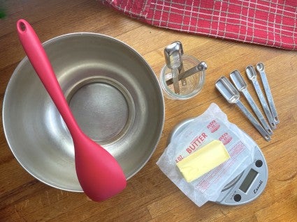 bowl, mixing spoon, scale with butter, and measuring spoons on a wooden table top.