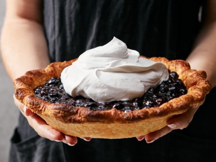 A baker holding a fresh blueberry pie topped with whipped cream