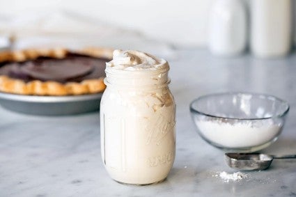 A mason jar of whipped cream with pumpkin pie in the background next to a bowl of 