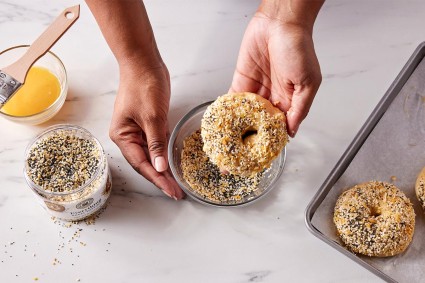 A baker dipping an egg washed bagel into a shallow bowl of seeds