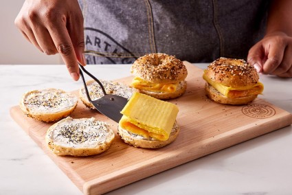 A baker placing an egg packet on an Ultimate Sandwich Bagel topped with a schmear of cream cheese