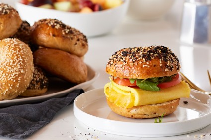 An Ultimate Sandwich Bagel filled with eggs, cheese, and tomatoes next to a platter of bagels