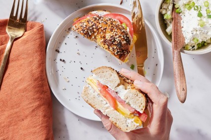 A person's hands holding half a bagel sandwich with tomato, as if about to bite into it