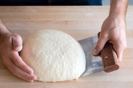 Hands kneading dough on a table with the aid of a bench knife.