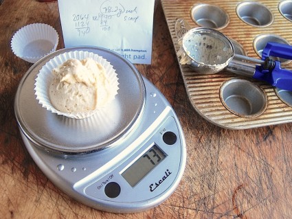 Scale with a paper liner of muffin batter on the platform, muffin pan and muffin scoop in the background.