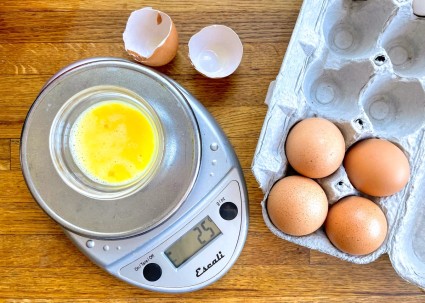 Braten egg in a small bowl set on a digital scale platform; carton of eggs and broken egg shell alongside.