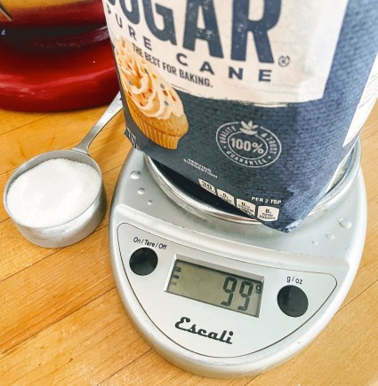 Bag of sugar on a digital scale, measuring cup of sugar alongside.