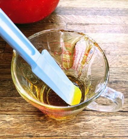 Glass measuring cup with honey in the bottom and smeared around the inside, silicone scraper in the cup.