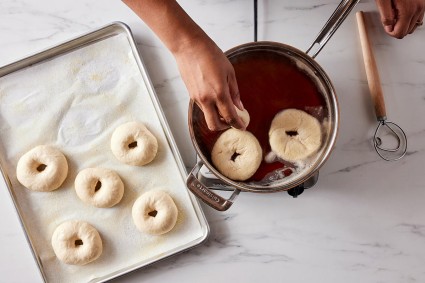 Bagels being placed in boiling water with hands, with dough whisk also on the counter nearby