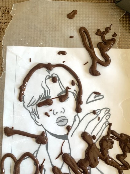 Chocolate icing blobbed all over a stencil drawing of a man's face, in the course of trying to trace the stencil.
