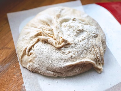 Bread dough turned out of a banneton onto a piece of parchment, totally deflated and looking like a limp balloon.