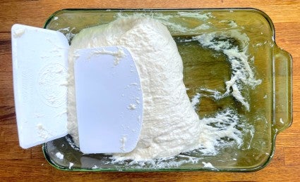 Slack Pan de Cristal dough in a green Pyrex casserole dish with bowl scrapers