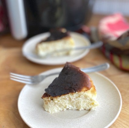 Wedge of Basque-style cheesecake on a plate with a fork, another plated cheesecake in the background, air fryer behind that.