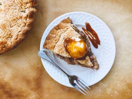 Piece of streusel-topped apple pie with vanilla ice cream and caramel sauce, on a white plate on a piece of golden-brown parchment.