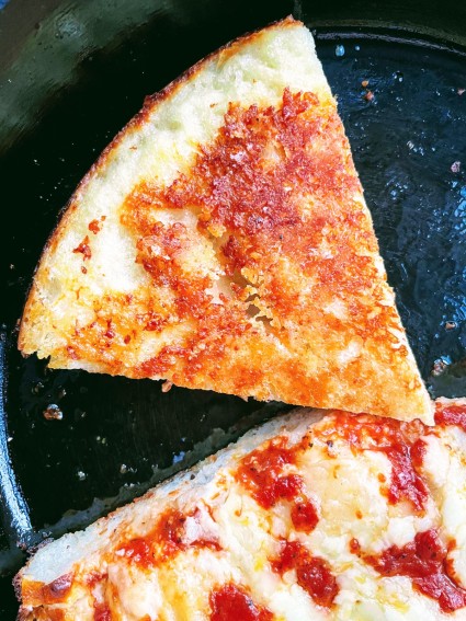 Two pieces of pizza in a cast iron skillet, one turned bottom-up to show crispy cheese on the bottom crust.