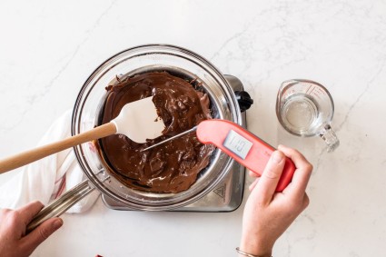 A baker taking the temperature of chopped chocolate melting over a water bath