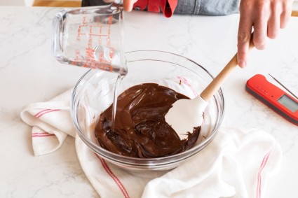 A baker adding warm corn syrup to a bowl of melted chocolate