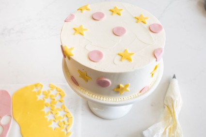 A white layer cake decorated with pink polka dots and yellow stars made from modeling chocolate