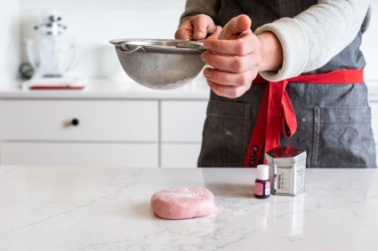 A baker sifting confectioners' sugar over the top of a disk of pink modeling chocolate