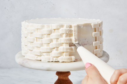 Hands piping a basket weave design on a cake with white frosting
