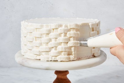 Hands piping a basket weave design on a cake with white frosting