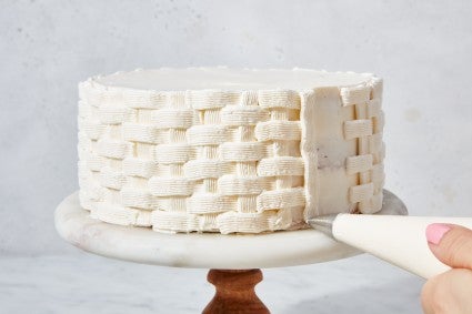 Hands piping a basket weave design on a cake with white frosting