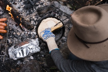 Baker using tongs to prepare pizza crust in a cast iron skillet over a campfire