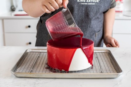 A baker pouring raspberry mirror glaze over a frosted, chilled cake