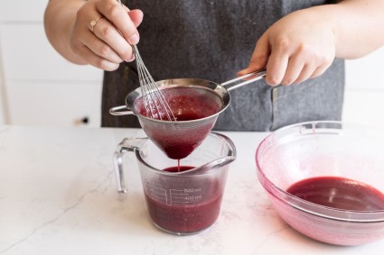 A baker straining blueberry mirror glaze into a measuring cup