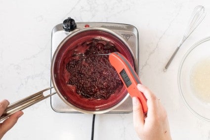 A baker testing the temperature of a pot of berry mirror glaze on an induction burner