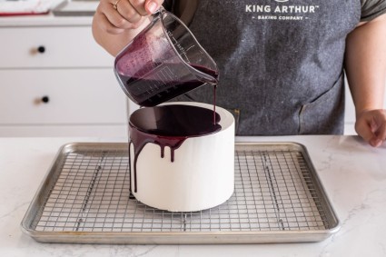 A baker pouring blueberry glaze over the top of a cake with drips