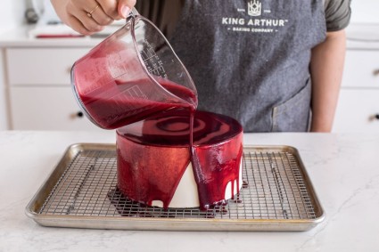 A baker pouring berry mirror glaze over a frosted cake