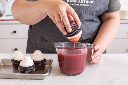 A baker dipping a frosted cupcake into a measuring cup of strawberry glaze