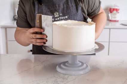 A baker smoothing the frosting on the sides of a cake using a bench knife