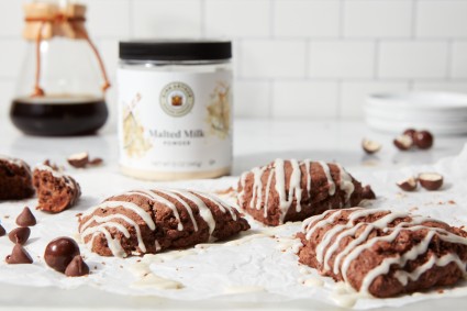 Chocolate scones drizzled with glaze on a piece of parchment in front of a jar of malted milk powder