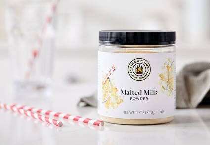 A jar of malted milk powder on a kitchen table next to two straws