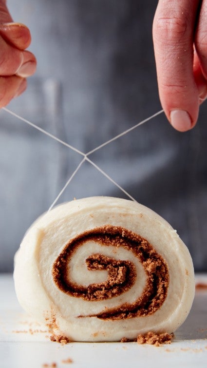 Log of cinnamon roll dough being cut into slices using dental floss.