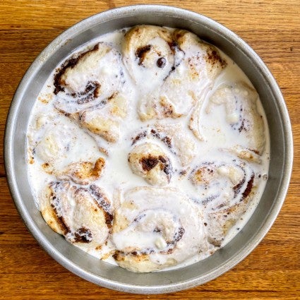 8" round pan of risen cinnamon rolls, soaked in heavy cream and ready to bake.