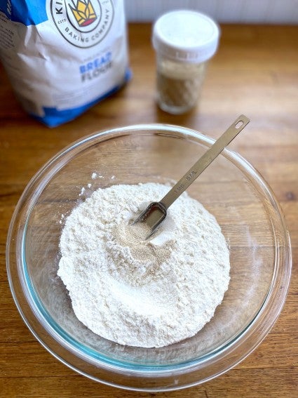 Instant yeast spooned into a bowl of flour, bread flour bag in the background