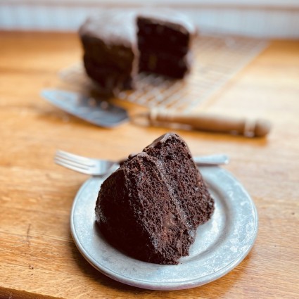 Thick wedge of chocolate-frosted chocolate cake on a pewter-type plate, whole cake in the background.