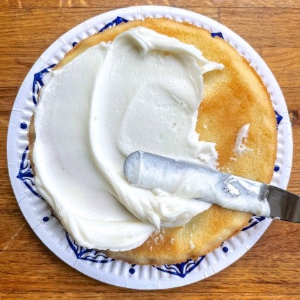 6-inch round yellow cake being frosted with white frosting using an offset spatula.