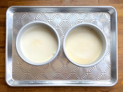Two 6" cake pans with yellow cake batter set on a half-sheet pan, ready to bake.