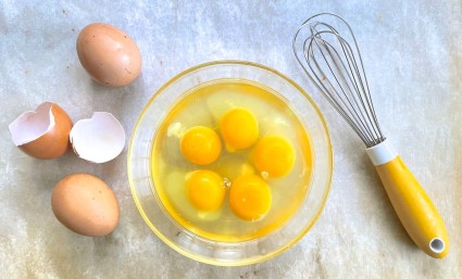 Five eggs cracked into a bowl, broken egg shells and a yellow-handled whisk on the side. on the side