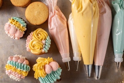 A baking sheet with pastry bags filled with different color frostings and cupcakes