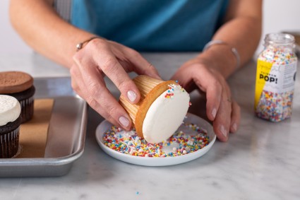 A baker rolling the sides of a flat-topped cupcake in naturally colored sprinkles