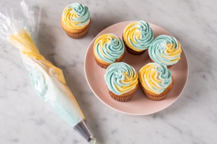 A plate of cupcakes frosted with two-tone frosting (blue and yellow) next to a piping bag with multicolored frosting